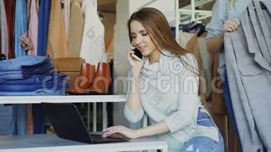 服装精品店的女老板正在用<strong>手机</strong>和笔记本电脑打字。 她的<strong>助手</strong>正在接近她，而且